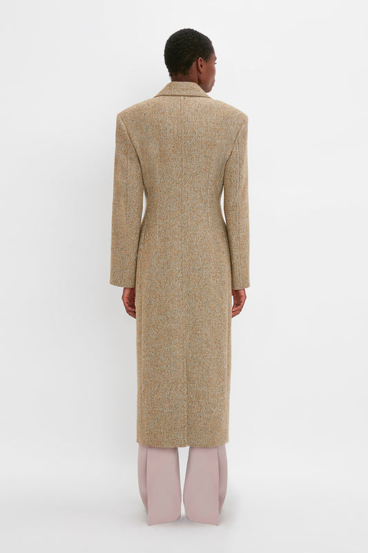 A person with short hair wearing a long, beige Victoria Beckham Exclusive Waisted Tailored Coat In Flax and light pink pants stands against a plain white background, facing away from the camera.