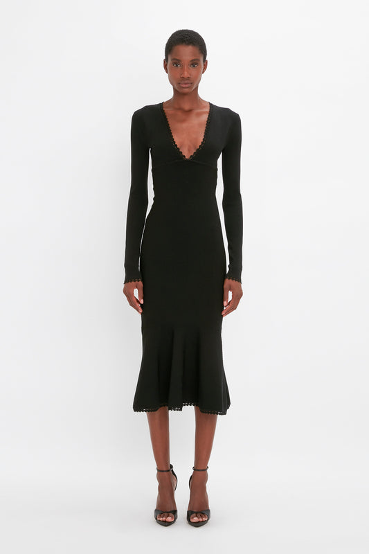 A person stands against a plain white background, wearing a VB Body Long Sleeve V Neck Dress In Black by Victoria Beckham with a plunging V-neckline and flared hem, paired with black strappy heels.