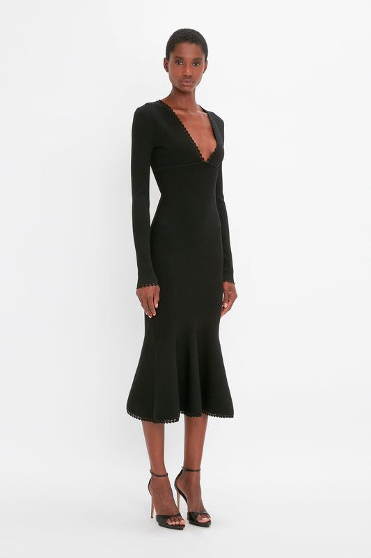 A person is standing against a white background wearing the VB Body Long Sleeve V Neck Dress in black by Victoria Beckham with a plunging V-neck, calf-length ruffled hem, and black high-heeled sandals.
