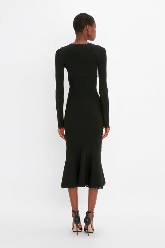 Person in a fitted Victoria Beckham VB Body Long Sleeve V Neck Dress In Black and high-heeled sandals is facing away, standing against a plain white background.