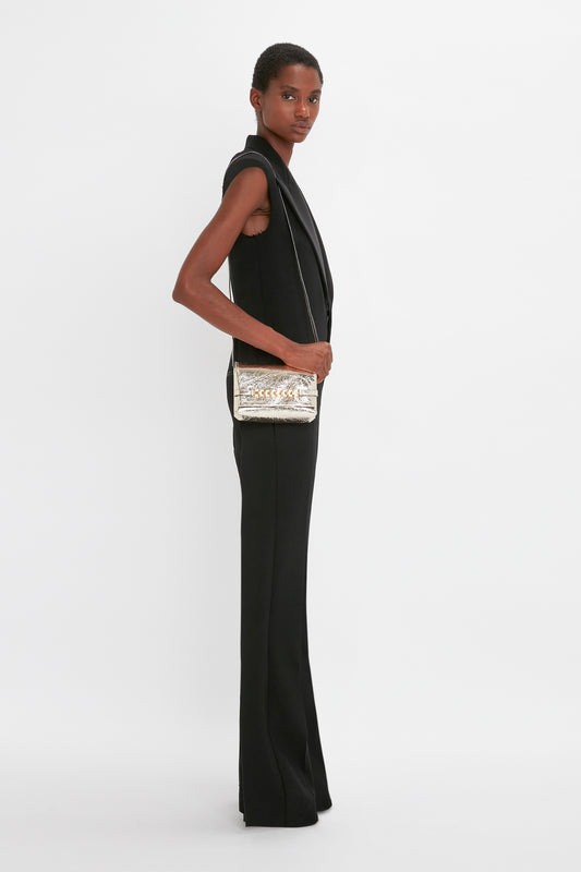 A woman in a sleek black sleeveless jumpsuit, standing sideways, looking over her shoulder, holding a Victoria Beckham Mini Chain Pouch With Long Strap In Gold Leather.