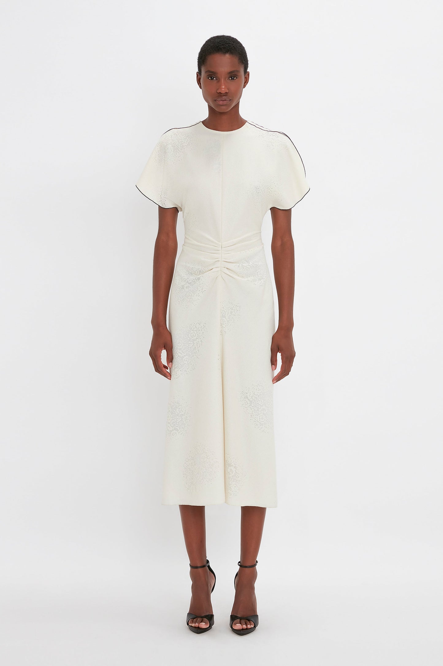 A woman stands facing forward, wearing a Victoria Beckham white embroidered gathered waist midi dress in cream with short sleeves and black strappy heels, on a plain white background.