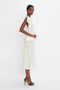 A person stands against a plain white background, wearing a sleeveless off-white dress and black high heels. They are holding an Exclusive Wallet On Chain In Ivory Croc-Effect Leather that evokes Victoria Beckham's chic style.