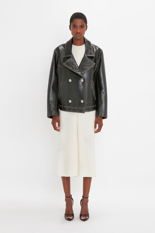 A person is standing against a white background, wearing an Oversized Leather Jacket In Black by Victoria Beckham with a distressed finish over a knee-length white dress and black ankle-strap sandals.