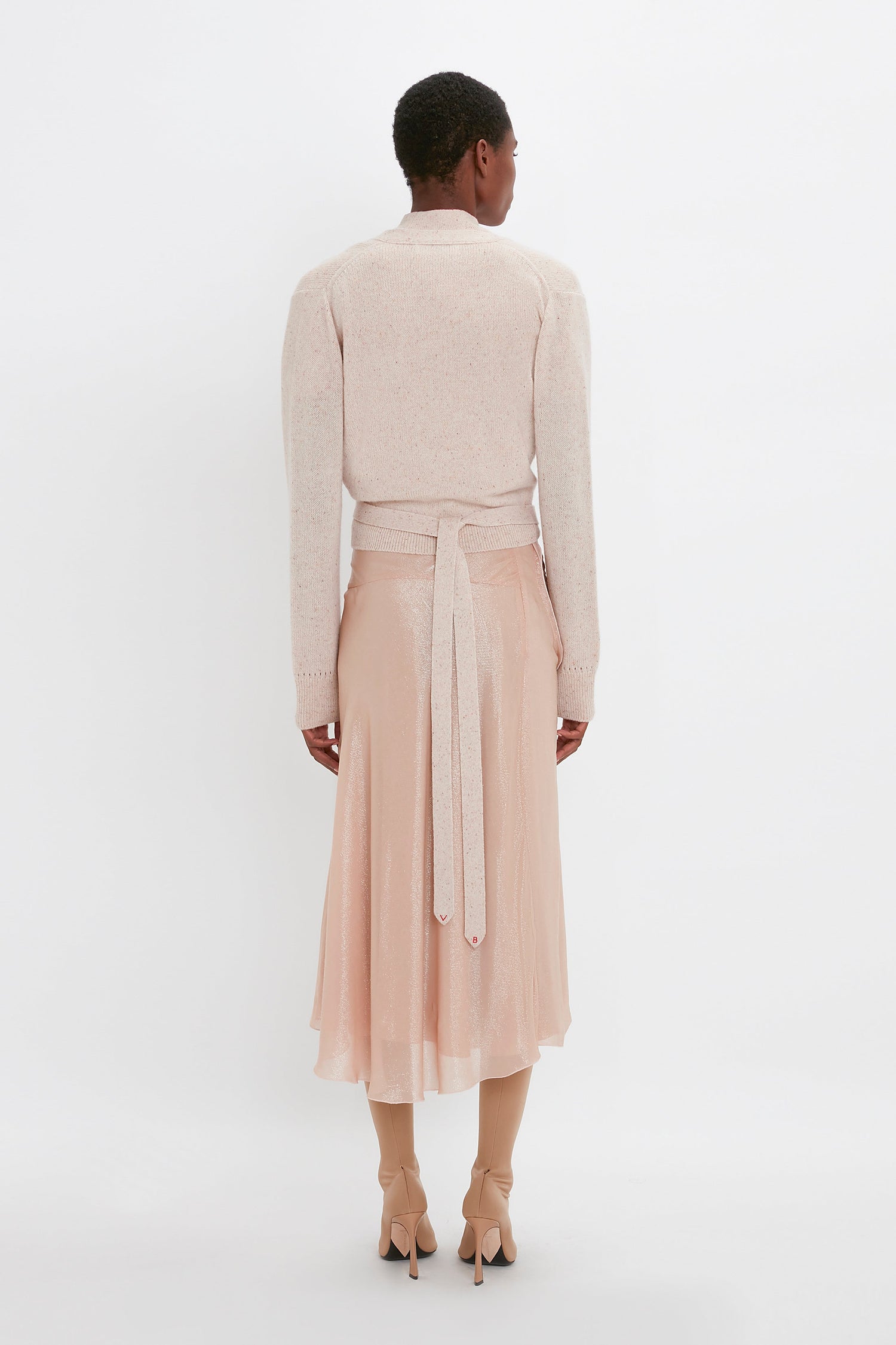 Person standing with their back to the camera, wearing a Victoria Beckham Wrap Front Cardigan In Nougat made of lightweight cashmere and a light peach-colored skirt with a wrap front detail, along with beige high-heeled shoes. The background is white, highlighting the versatility of this stylish ensemble.