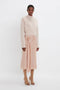 A person stands against a plain white background, wearing the Victoria Beckham Wrap Front Cardigan In Nougat, a pale peach mid-length skirt with a floral embellishment, and beige heeled open-toe shoes. The versatile styling captures an effortless elegance.