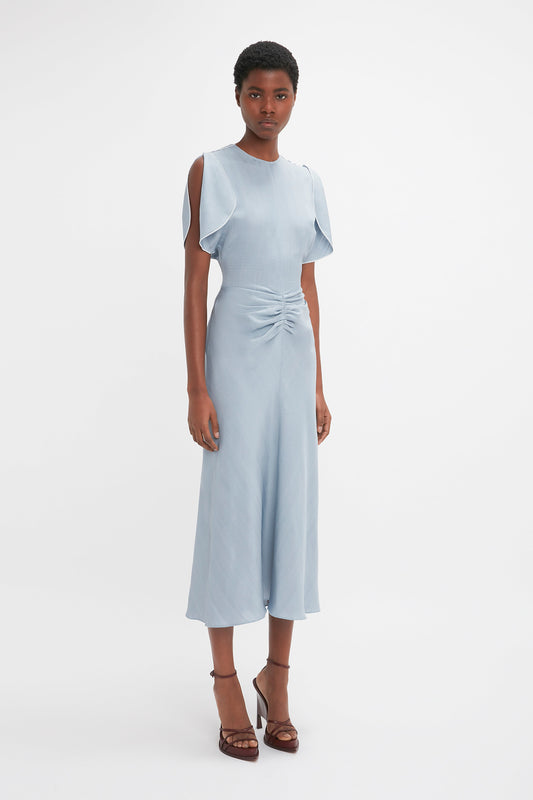 A woman in a Exclusive Gathered Waist Midi Dress In Pebble by Victoria Beckham with ruffle sleeves and a front twist stands against a white background, wearing brown heels.