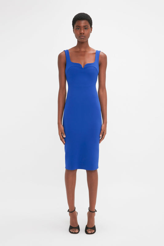 A person stands facing the camera, wearing a Victoria Beckham Sleeveless Fitted T-Shirt Dress In Palace Blue with a sweetheart neckline and black high-heeled shoes.