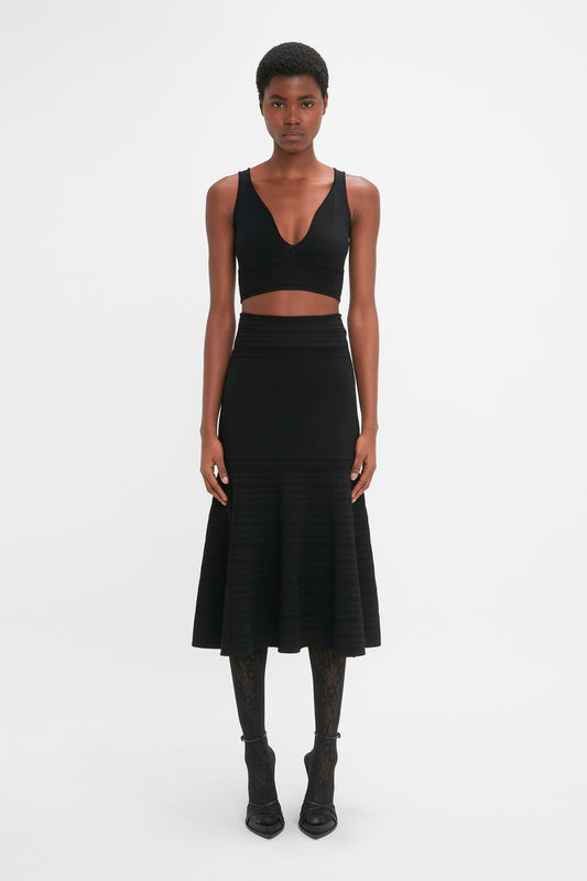 A person stands facing forward, wearing a black Victoria Beckham Frame Detail Sleeveless Top In Black with contrasting stitch details, a black midi skirt, and black heeled shoes. The background is plain white.
