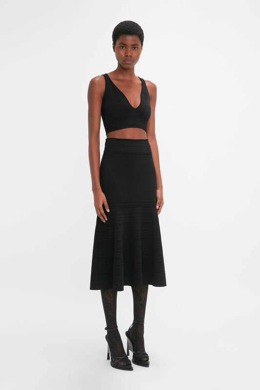 Person wearing a Victoria Beckham Frame Detail Sleeveless Top In Black and a black high-waisted skirt with a flared hem, standing against a plain white background.