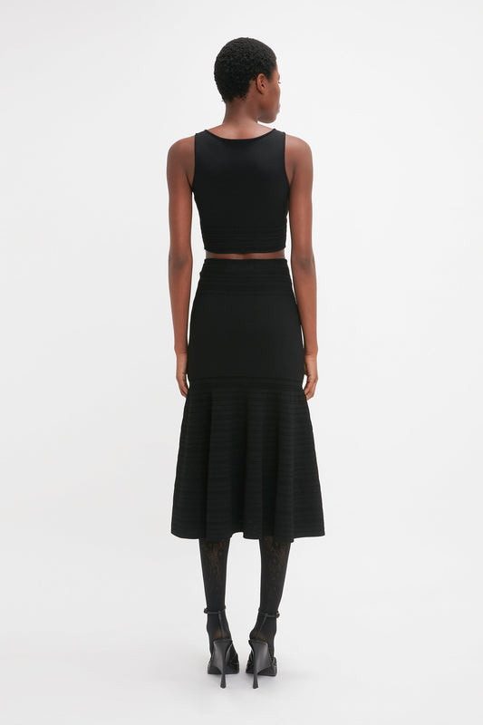 Person standing against a white background, wearing a black Frame Detail Sleeveless Top In Black by Victoria Beckham and a black midi skirt with contrasting stitch details, coupled with black patterned tights and high-heeled shoes. The body's sculpting fit is emphasized as the individual faces away from the camera.