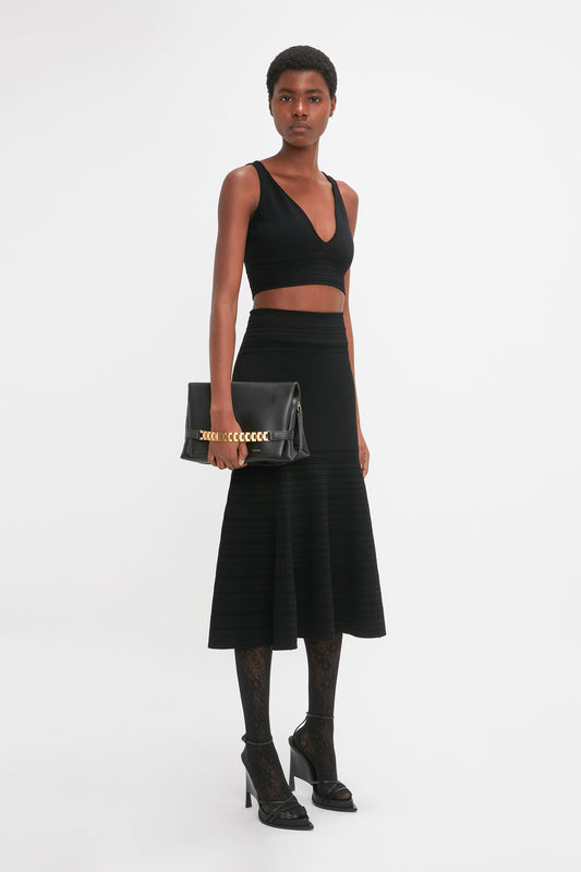 A person stands against a white background wearing a Victoria Beckham Frame Detail Sleeveless Top In Black, a black high-waisted skirt, black heels, and patterned tights, holding a black clutch with gold chain accents.