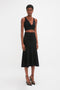 A person with short hair is wearing a black sleeveless crop top and a waist-defining Fit And Flare Midi Skirt In Black from Victoria Beckham made from mid-weight stretch knit, paired with black high-heeled sandals, posing against a plain white background.