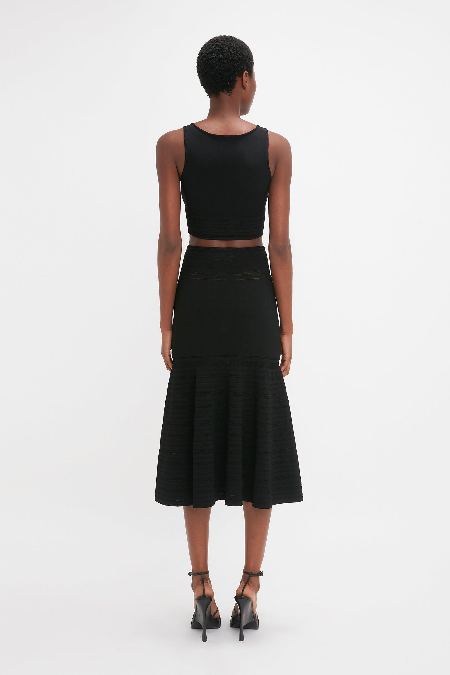 Person standing with their back to the camera, wearing a black sleeveless crop top, Victoria Beckham Fit And Flare Midi Skirt In Black, and black high-heeled shoes.