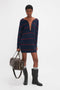 Person standing in a studio wearing the Victoria Beckham Frame Detail Jumper Dress In Navy-Red, black boots, and holding a brown leather bag. The mid-weight knit garment features a directional curved neckline, adding a touch of modern elegance.