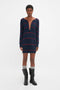 A person stands facing forward wearing a navy blue, Frame Detail Jumper Dress In Navy-Red by Victoria Beckham with red horizontal stripes and a directional curved neckline. They have short hair and are dressed in black knee-high boots with gray socks showing above the boots.