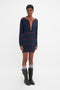 A person stands facing forward wearing a Frame Detail Jumper Dress In Navy-Red by Victoria Beckham with a deep directional curved neckline, gray knee-high socks, and black boots. The background is plain white.