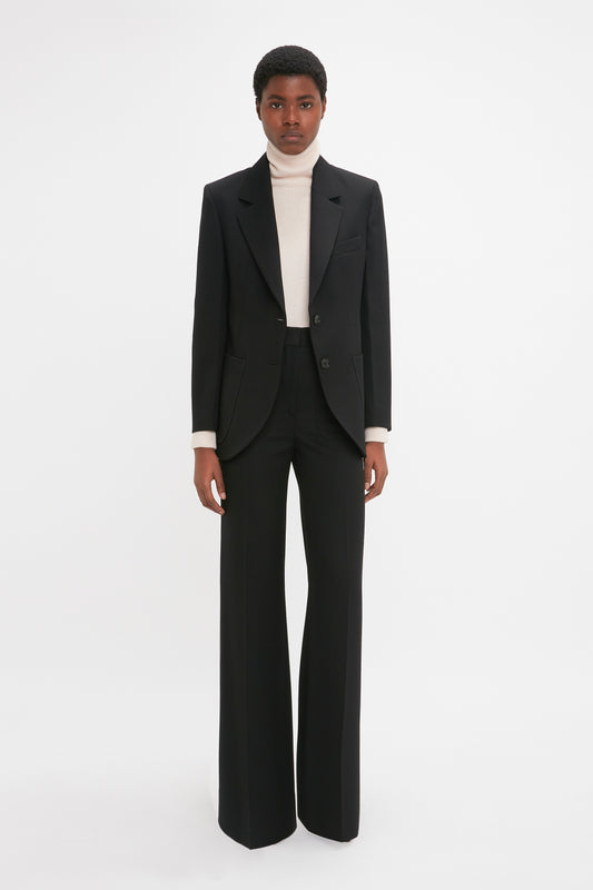 Person standing and facing forward, wearing a Victoria Beckham Patch Pocket Jacket In Black with trousers, paired with a white turtleneck, posed against a plain white background.