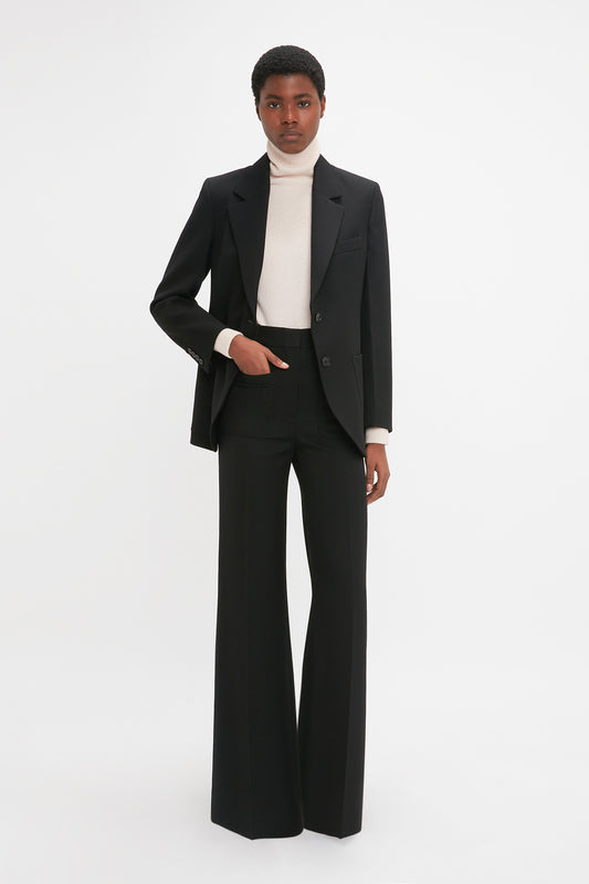 Person dressed in a Victoria Beckham Patch Pocket Jacket In Black, white turtleneck, and black wide-leg trousers standing against a plain white background.