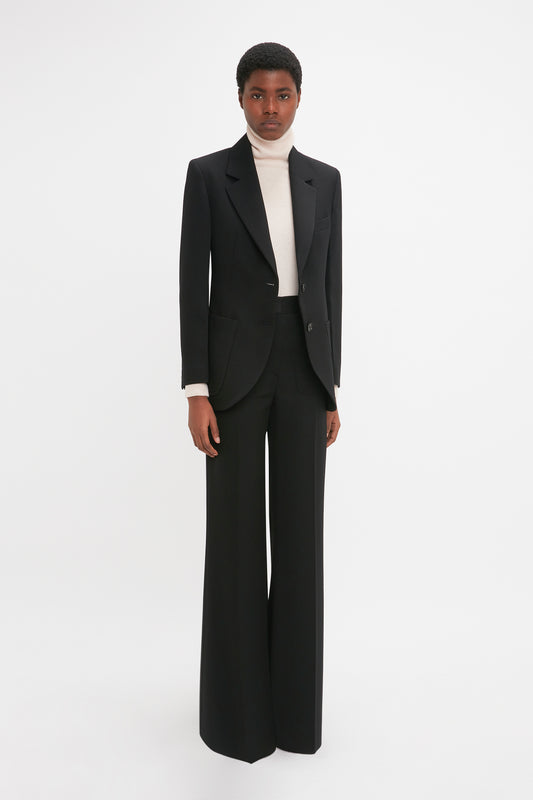 A person wearing a Victoria Beckham Patch Pocket Jacket In Black over a white turtleneck with matching black wide-leg pants stands against a plain white background.