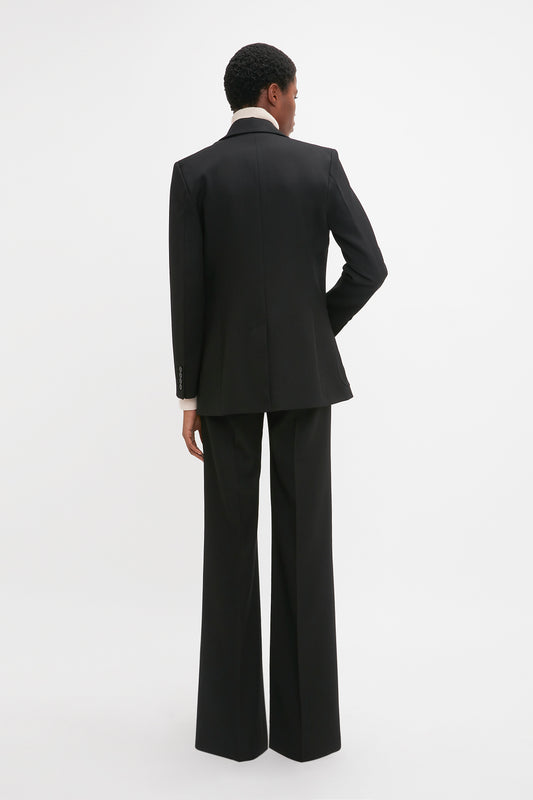 A person with short hair is standing and facing away, wearing the Victoria Beckham Patch Pocket Jacket In Black with flared pants and a white shirt, exuding a tailored fit against a plain white background.