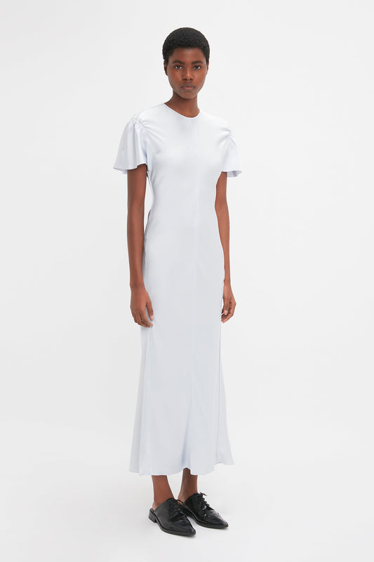 A person stands wearing a light blue, short-sleeved, elegant Gathered Sleeve Midi Dress In Ice by Victoria Beckham and black shoes against a plain white background.
