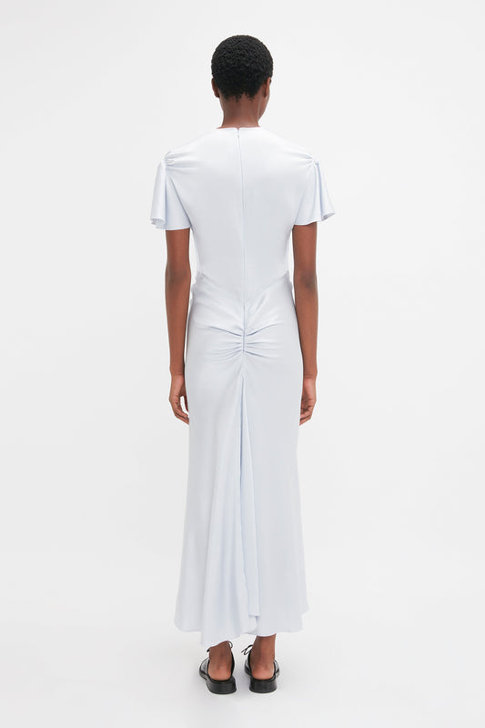 A person stands facing away, wearing the Victoria Beckham Gathered Sleeve Midi Dress In Ice in light blue with short sleeves and ruched detailing on the back. The ankle-length gown features a subtle godet insert and is paired perfectly with black shoes, all against a plain white background.
