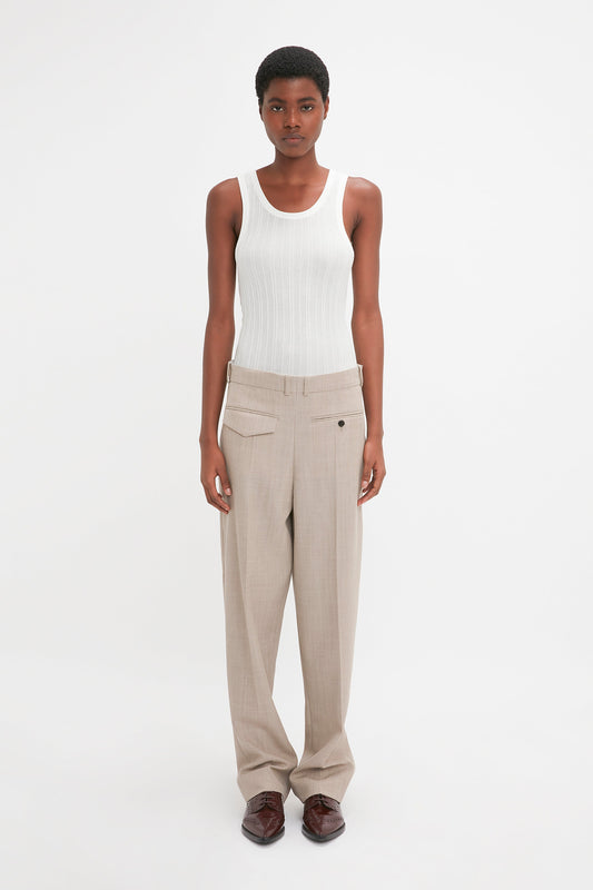 A person is standing against a plain white background wearing a white sleeveless top and the Reverse Front Trouser In Sesame by Victoria Beckham, with hands relaxed by their sides, showcasing a contemporary silhouette.