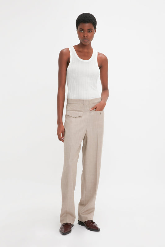 Person with short hair wearing a white tank top, beige trousers, and brown shoes, standing against a plain white background with one hand in their pocket. The Fine Knit Vertical Stripe Tank In White gives a Victoria Beckham-inspired minimalist vibe.
