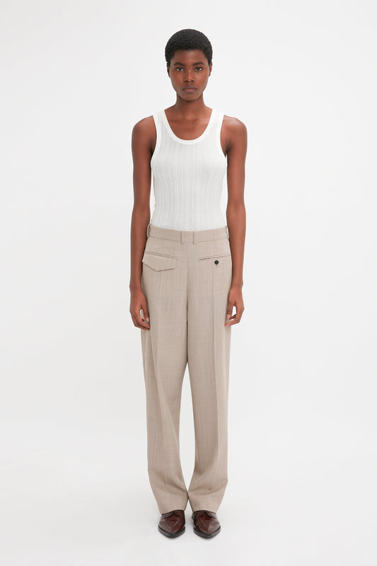 A person stands facing the camera against a plain white background, wearing the Fine Knit Vertical Stripe Tank In White by Victoria Beckham and beige high-waisted pants. They have short hair and are seen from the front, reminiscent of Victoria Beckham's minimalist style.