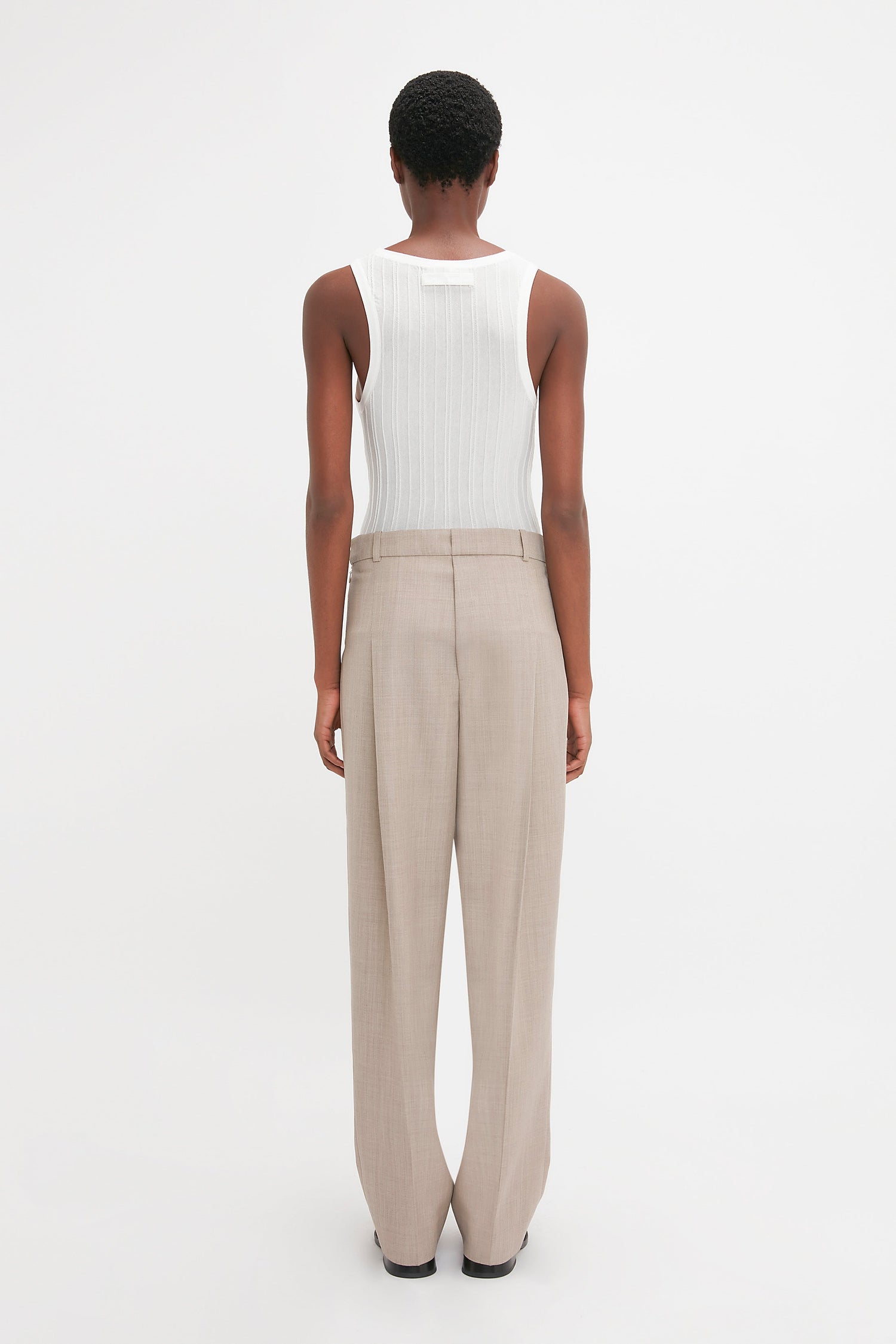 Person facing away from the camera, wearing a Victoria Beckham Fine Knit Vertical Stripe Tank In White and beige trousers, against a plain white background.