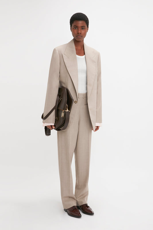 A person with short hair is wearing a Darted Sleeve Tailored Jacket In Sesame by Victoria Beckham over a white shirt. They are holding a brown leather bag and standing against a plain white background, showcasing contemporary detailing.