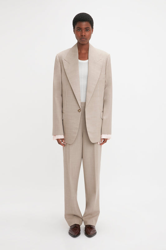 A person stands against a plain white background wearing the Victoria Beckham Darted Sleeve Tailored Jacket In Sesame with contemporary detailing and a single-button closure, a white inner shirt, and brown shoes. They have a neutral expression and short hair.