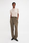 A person stands against a plain background, wearing the Victoria Beckham Short Sleeve Top In Nougat with contrasting trim and high-waisted olive green pants, black shoes on their feet, hands in pockets, and looking forward.