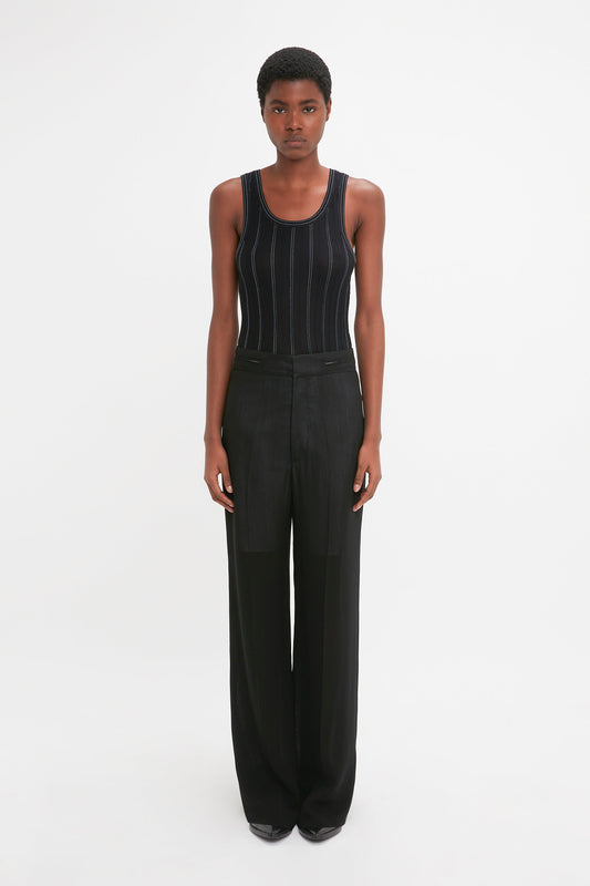 A person stands against a plain white background wearing a sleek Victoria Beckham black sleeveless Fine Knit Vertical Stripe Tank In Black-Blue and black wide-leg pants.