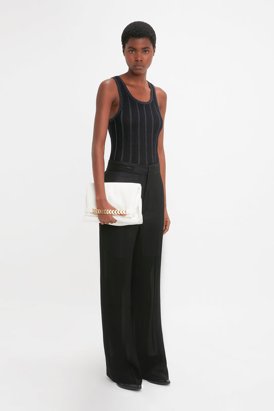 A person with short hair wears a black Fine Knit Vertical Stripe Tank In Black-Blue and long black pants, holding a white clutch. They stand against a plain white background, evoking the minimalist elegance of Victoria Beckham's designs.