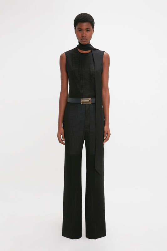 Person standing against a plain white background, wearing a Sleeveless Tie Neck Top In Black by Victoria Beckham and high-waisted, wide-leg black trousers made of crepe de chine silk, secured around the waist by a belt with a gold buckle.