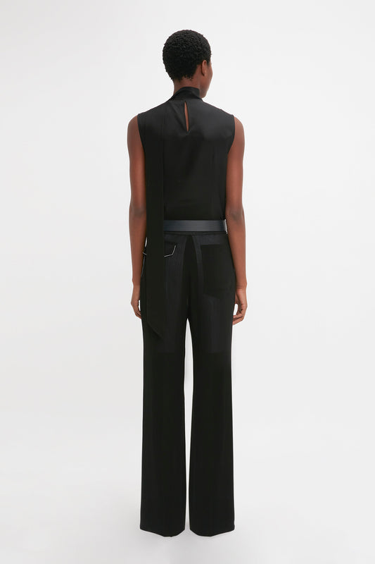 Person stands facing away from the camera, wearing the Victoria Beckham Sleeveless Tie Neck Top In Black paired with loose-fitting black trousers. The outfit includes a wide black belt with a silver clasp.