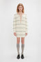 A person with medium-length wavy hair is wearing an off-white, long-sleeve, striped Frame Detail Jumper Dress In Natural-Navy by Victoria Beckham with a deep V-neck, gray knee-high socks, and black shoes. This mid-weight knit piece channels the SS24 runway vibes perfectly.