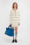 A person with long hair wears a cream-colored, striped Frame Detail Jumper Dress In Natural-Navy by Victoria Beckham with a deep V-neck, gray knee-high socks, and black shoes. They hold a large blue bag with both hands, reminiscent of the chic SS24 runway styles.