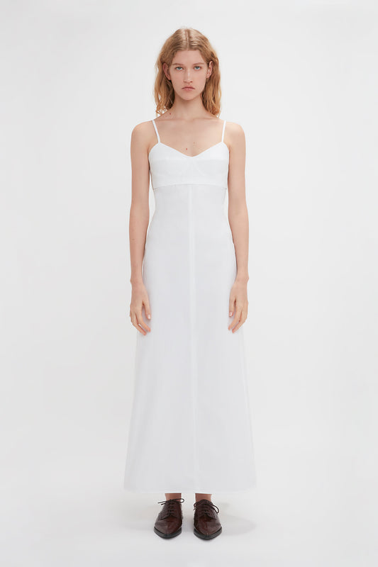 A person with blonde hair is standing in a Victoria Beckham Cami Fit And Flare Midi In White with adjustable straps and brown shoes against a white background, facing the camera.