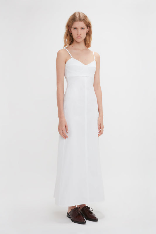 A woman in a long, white spaghetti-strap summer dress called the Cami Fit And Flare Midi In White by Victoria Beckham stands against a plain white background. She is wearing dark brown oxford shoes.