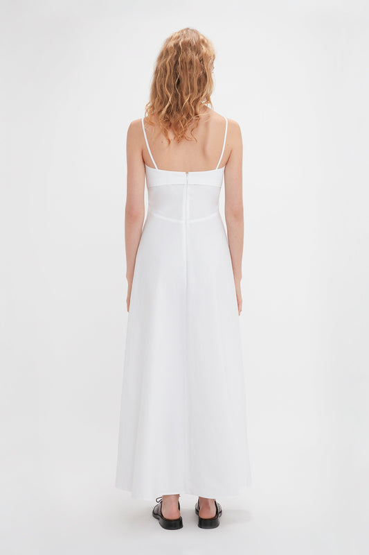 A person with wavy blonde hair is seen from the back, wearing the Victoria Beckham Cami Fit And Flare Midi In White with adjustable straps and black shoes. Made from breathable stretch cotton, the dress has a zipper running down the back.