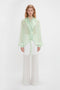 A person in a Victoria Beckham Romantic Blouse In Jade with diaphanous blouson sleeves and sheer white pants stands against a plain white background.