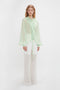 A person stands wearing a Romantic Blouse In Jade by Victoria Beckham with diaphanous blouson sleeves and semi-transparent white pants against a plain white background.