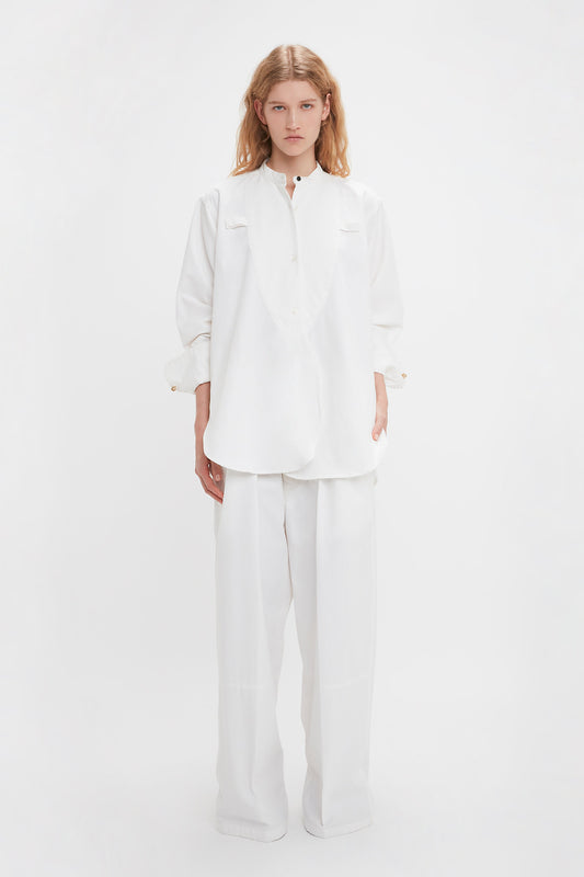 A person with long, light-colored hair stands against a white backdrop, wearing all-white oversized clothing, including a relaxed fit 100% cotton Bib-Front Tuxedo Shirt In Washed White by Victoria Beckham and wide-leg pants.