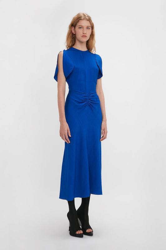 A woman in a Victoria Beckham Gathered Waist Midi Dress in Palace Blue, knee-length with cap sleeves and a twisted front detail, paired with black open-toe heels, standing against a white background.