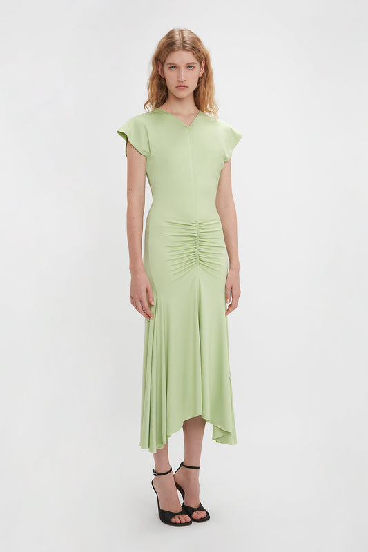 A woman stands against a white background, wearing a light pistachio, mid-length Sleeveless Rouched Jersey Dress by Victoria Beckham with black heels.