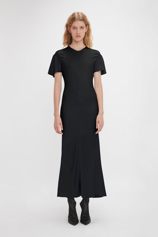 A person with long hair is wearing a black Gathered Sleeve Midi Dress In Black by Victoria Beckham and black lace-up shoes, standing against a white background.