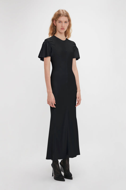 A woman with long hair is standing and wearing the Victoria Beckham Gathered Sleeve Midi Dress In Black paired with black ankle boots. She is facing the camera with a neutral expression against a plain white background.